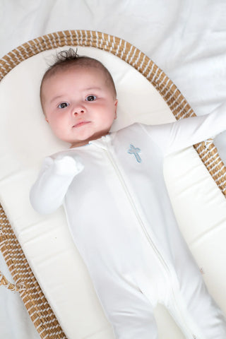 Baby in a white outfit with a blue cross symbol, lying in a Moses basket.