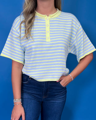 A woman wearing a light blue and yellow striped short-sleeve henley top with a yellow collar and button placket, paired with dark blue jeans