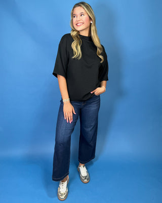 A woman modeling a black short-sleeve mock neck t-shirt with dark blue wide-leg jeans and white sneakers