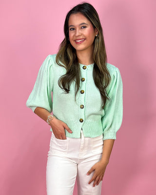 Woman wearing a light green cardigan with gold buttons against a pink background