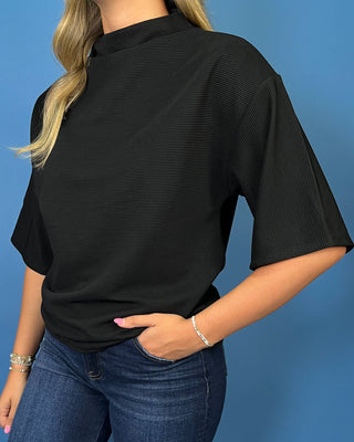 A woman wearing a black short-sleeve top with a textured, ribbed fabric and a mock neck, paired with blue jeans