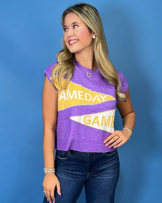 A smiling woman with blonde hair wearing a purple knit sweater with yellow and white graphic text that reads 'GAMEDAY' and blue jeans.