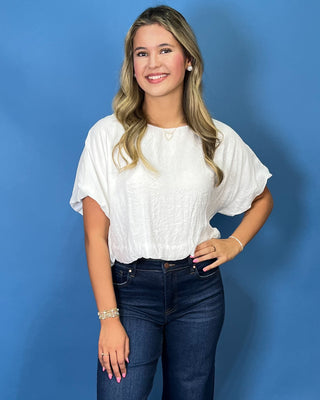 A woman with blonde hair wearing a white textured short-sleeve blouse and dark blue jeans