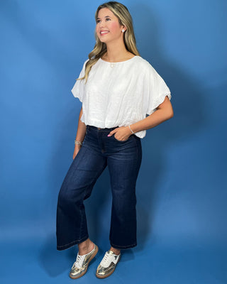 A woman modeling a white short-sleeve top with flutter sleeves, paired with dark blue wide-leg jeans and metallic gold sneakers