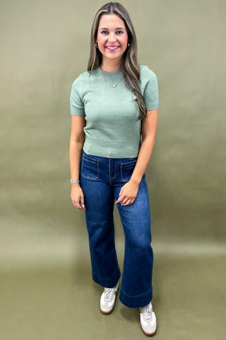 Woman wearing a green sweater and blue jeans against a beige background