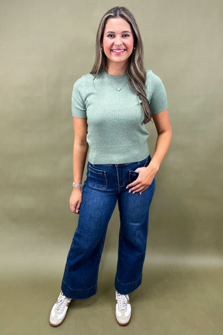 Woman wearing a green sweater and blue jeans against a plain background