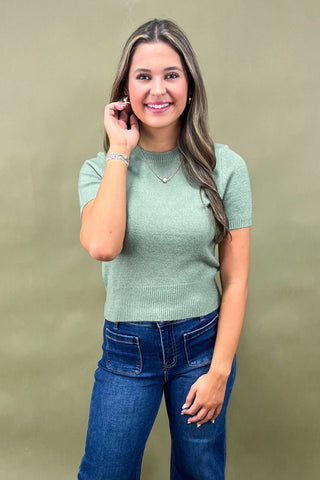 Woman wearing a green sweater and blue jeans against a beige background