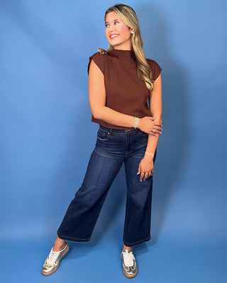 A woman modeling a brown ribbed mock neck top with cap sleeves and a decorative gold-toned embellishment on the shoulder