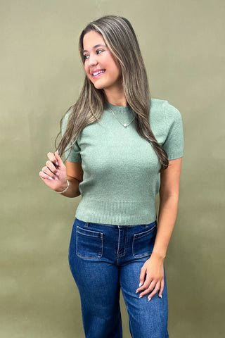 Woman wearing a green sweater and blue jeans against a beige background