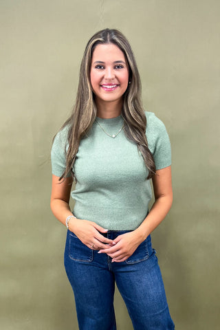 Woman wearing a green sweater and blue jeans against a beige background