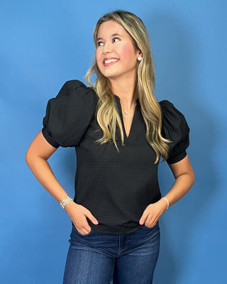 A woman modeling a black short-sleeve blouse with puffed shoulders and a V-neck, paired with dark blue jeans and white sneakers