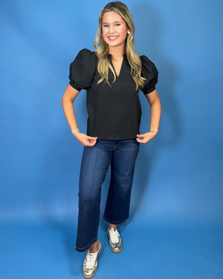 A woman modeling a black short-sleeve blouse with puffed shoulders and a V-neck, paired with dark blue jeans and white sneakers