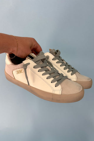 Beige sneakers with brown soles held by a hand against a light blue background
