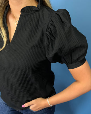 A close-up of a woman wearing a black textured short-sleeve blouse with a ruffled V-neckline and puff sleeves, paired with blue jeans