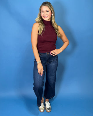 A woman modeling a sleeveless ribbed mock neck top in burgundy, paired with dark blue cropped jeans and white sneaker