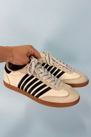 Beige sneakers with black stripes held by a hand against a light blue background