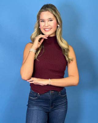 A woman modeling a sleeveless ribbed mock neck top in burgundy, paired with dark blue cropped jeans and white sneaker
