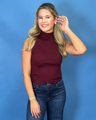 A woman modeling a sleeveless ribbed mock neck top in burgundy, paired with dark blue jeans
