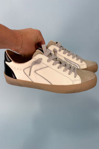 Pair of beige sneakers with black accents held by a hand against a light blue background