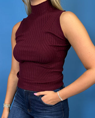 A woman modeling a sleeveless ribbed mock neck top in burgundy, paired with dark blue cropped jeans and white sneaker