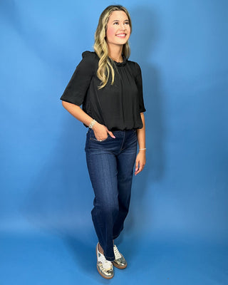 A woman modeling a black short-sleeve blouse with a gathered neckline, paired with dark blue jeans and white sneakers with gold accents,