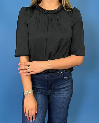 A woman wearing a black short-sleeve blouse with a gathered, scalloped ruffle neckline, paired with dark blue jeans