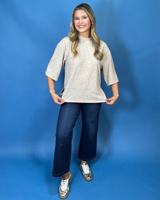 A woman modeling a light beige heathered short-sleeve top with flared bell sleeves, paired with dark blue wide-leg jeans and metallic silver sneakers