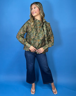 Olive green floral print long sleeve blouse with a ruffled neckline, statement bow detail, and warm brown and sage accents.