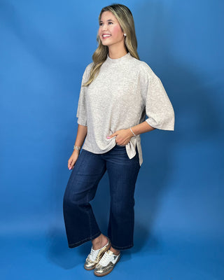 A woman modeling a light beige heathered short-sleeve top with elbow-length flared sleeves and a side tie detail, paired with dark blue jeans and metallic sneaker