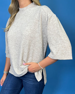 A woman wearing a light heather gray oversized short-sleeve top with a side split and tie detail, paired with blue jeans.