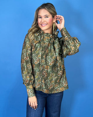 Olive green floral print long sleeve blouse with a ruffled neckline, statement bow detail, and warm brown and sage accents.
