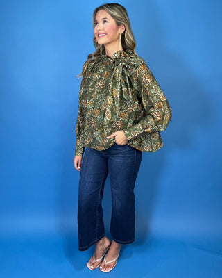 Olive green floral print long sleeve blouse with a ruffled neckline, statement bow detail, and warm brown and sage accents.