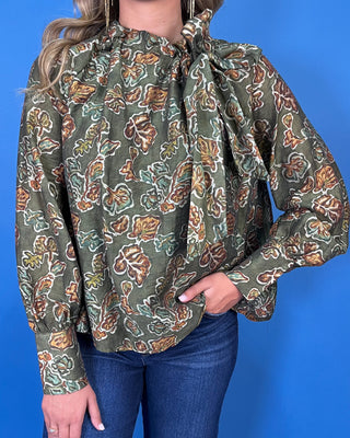 Olive green floral print long sleeve blouse with a ruffled neckline, statement bow detail, and warm brown and sage accents.