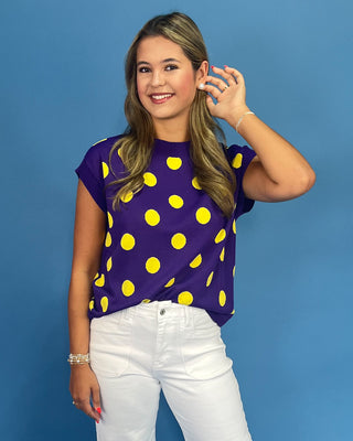 Woman wearing a purple top with yellow polka dots and white pants against a blue background