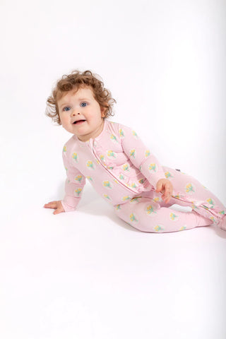 A baby with curly brown hair wearing a pink footed sleeper with a green and yellow pattern, lying on a white studio background.