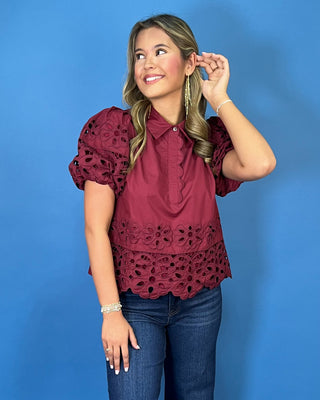 Burgundy eyelet blouse with statement puff sleeves, a collared neckline, scalloped trim, and intricate floral cutout details.