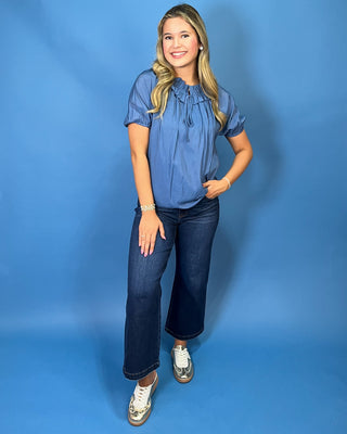 A woman modeling a light blue short-sleeve blouse with puff sleeves and a gathered neckline, paired with dark blue jeans and metallic sneakers