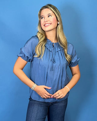 A woman modeling a light blue short-sleeve blouse with puff sleeves and a gathered neckline, paired with dark blue jeans and metallic sneakers