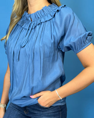 A woman wearing a blue short-sleeve blouse with a ruffled neckline and tie detail, paired with dark blue jeans