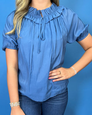 A woman wearing a blue short-sleeve blouse with a ruffled high neck and tie detail, paired with dark blue jeans