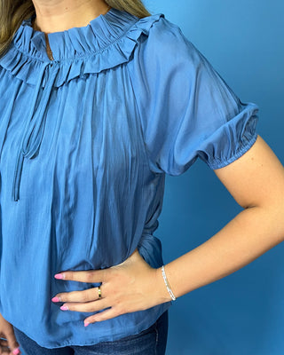 A woman wearing a dusty blue short-sleeve blouse with a ruffled neckline and drawstring details