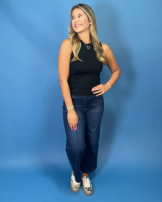 A woman modeling a black sleeveless ribbed tank top and dark blue cropped jeans with white sneakers