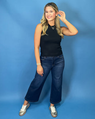 A woman modeling a black sleeveless ribbed tank top and dark blue wide-leg jeans with metallic silver sneakers
