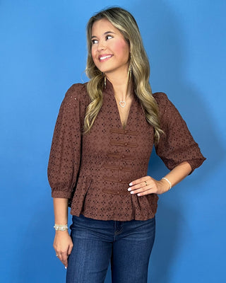 Chocolate brown eyelet peplum top with a V-neckline, decorative bow-and-button front details, and three-quarter puff sleeves.