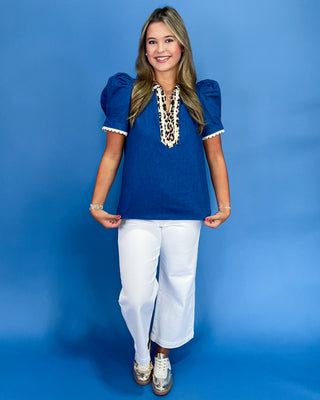 Woman wearing a blue top with lace details and white pants against a blue background