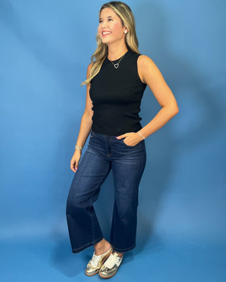 A woman modeling a black sleeveless ribbed tank top and dark blue cropped flare jeans