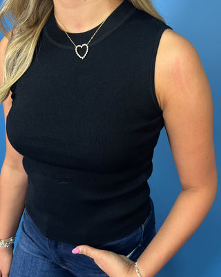 A woman wearing a black ribbed sleeveless mock neck top and blue jeans, accessorized with a small heart-shaped necklace