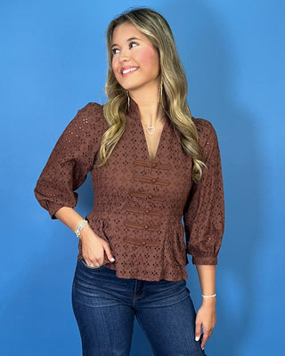 Chocolate brown eyelet peplum top with a V-neckline, decorative bow-and-button front details, and three-quarter puff sleeves.