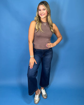 A woman posing in a sleeveless ribbed taupe-brown mock neck top and dark blue wide-leg jeans