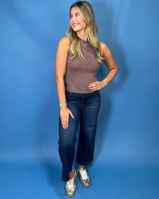 A woman posing in a brown ribbed sleeveless mock neck top, dark blue cropped jeans, and metallic silver sneakers 